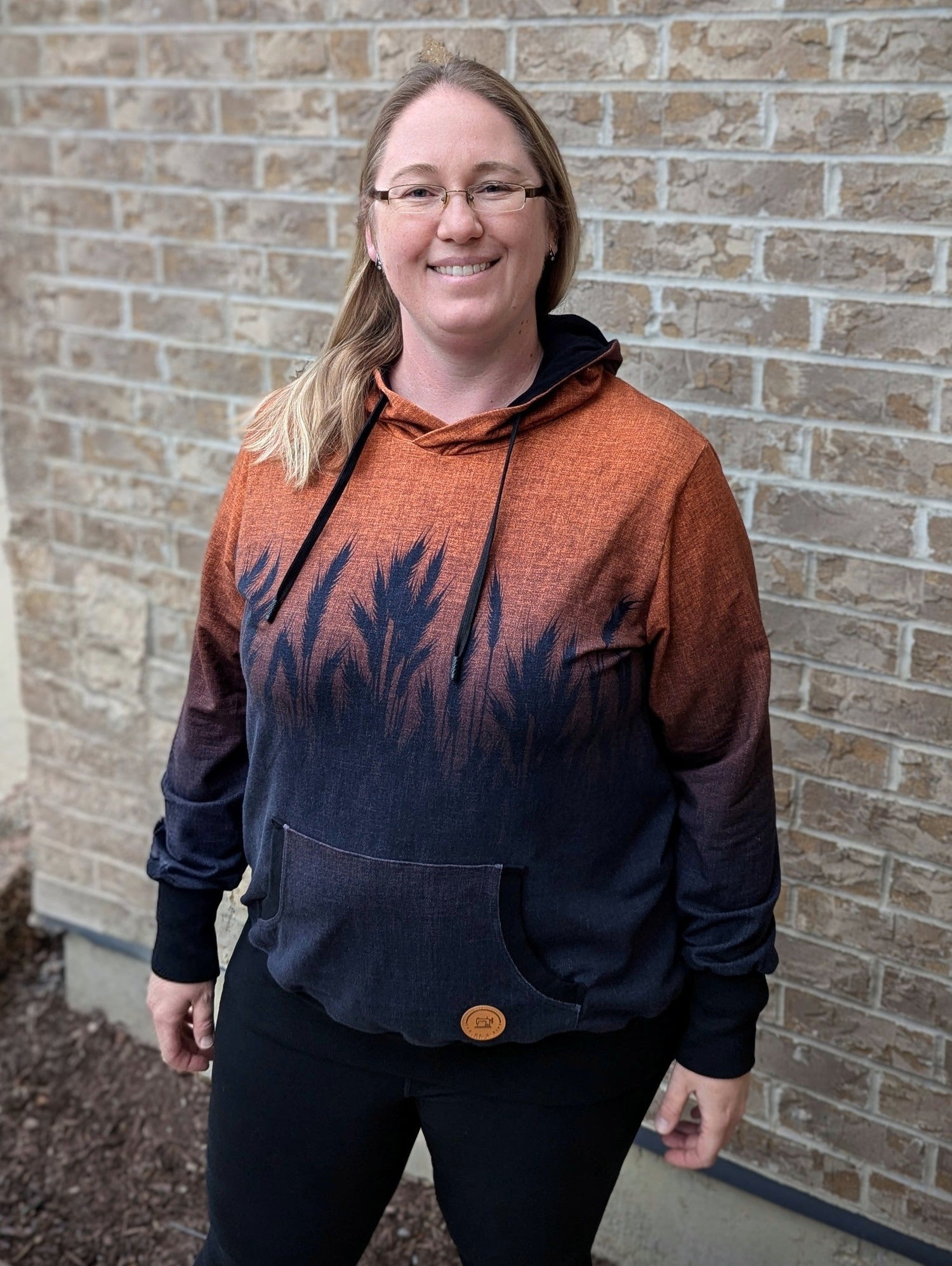 An adult female with blonde hair and glasses wearing a long-sleeved hoodie printed with a large black wheat design over an orange heathered background hoodie stands in front of a brick wall.