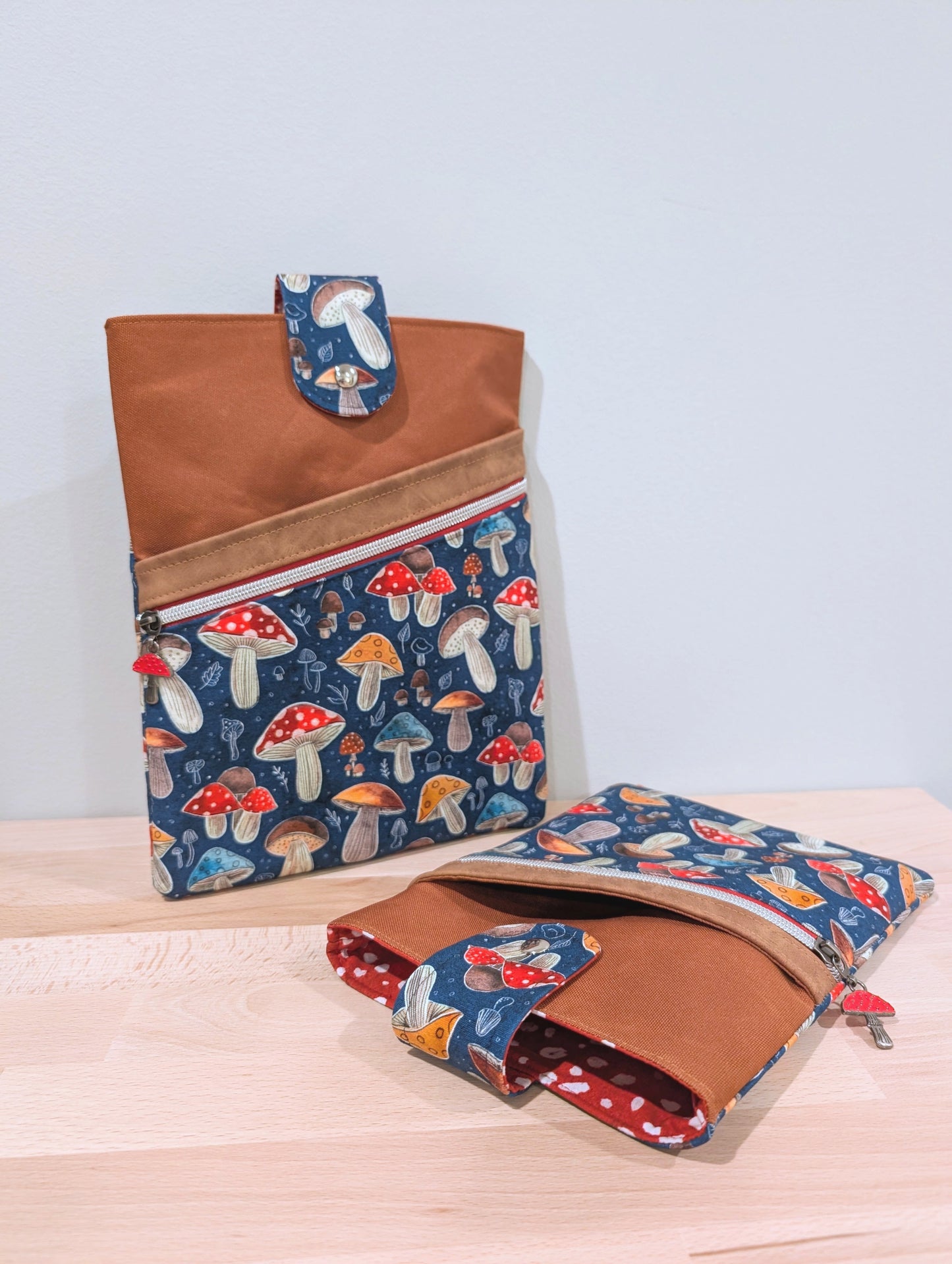 Two fabric sleeves of different sizes with brown, a diagonal red zipper with sliver teeth, a red mushroom zipper pull, and blue mushroom-print lower panels. The lining of the sleeve is a red background with white dots to match the mushroom caps.