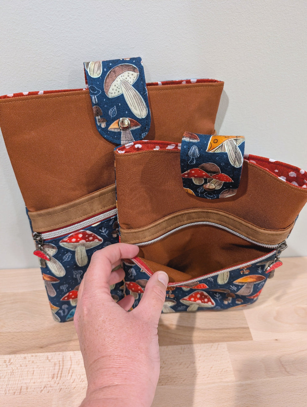 A close-up of a hand opening a one of two fabric sleeves of different sizes with brown, a diagonal red zipper with sliver teeth, a red mushroom zipper pull, and blue mushroom-print lower panels.